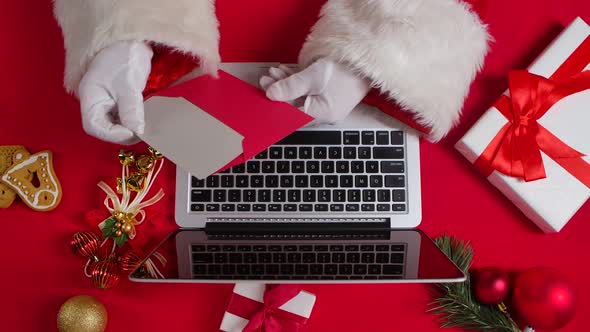 Top View Santa Hands in White Gloves are Typing on Keyboard Laptop By Red New Year Decorated Table alt