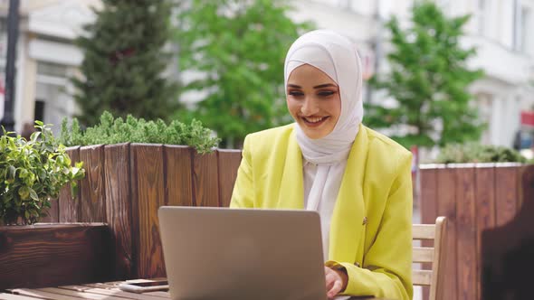 Smiling Young Muslim Woman Wearing Headscarf Sits in Cafe and Uses Laptop