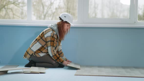 Female Construction Professional in Safety Helmet Lays Laminate Flooring Indoors During Renovation alt