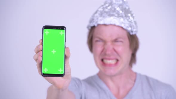 Face of Stressed Young Man with Tinfoil Hat Showing Phone and Looking Scared alt
