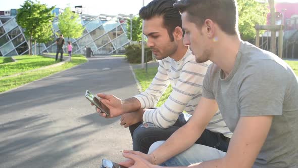 two young multi ethnic men outdoor sitting using smart phone having conversation alt