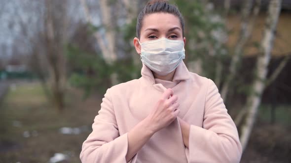 woman in medical mask wraps herself in a coat on the street alt