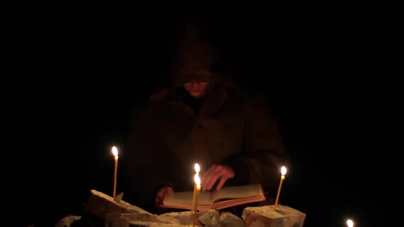 a Man in a Cloak with a Hood is Reading a Hot Book with Candles on the Floor alt