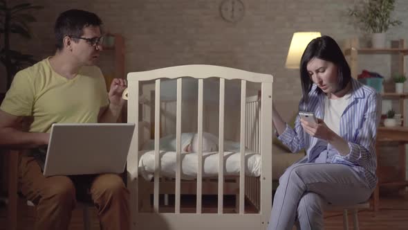 Young Parents Search the Internet and a Newborn in a Baby Bed alt