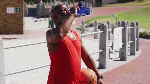 Senior african american man with basketball drinking water on the court near the beach alt