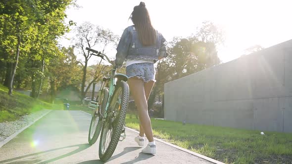 View from Back of Young Woman Walking on City Streets with Bicycle in Summer Day alt