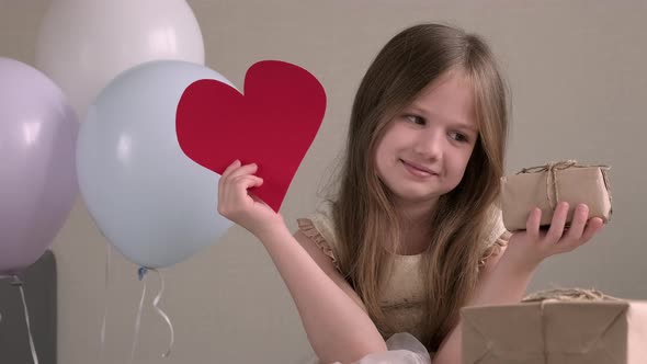 Little Girl Holding Big Red Paper Heart and Zero Waste Gift on Background of Air Balloons alt