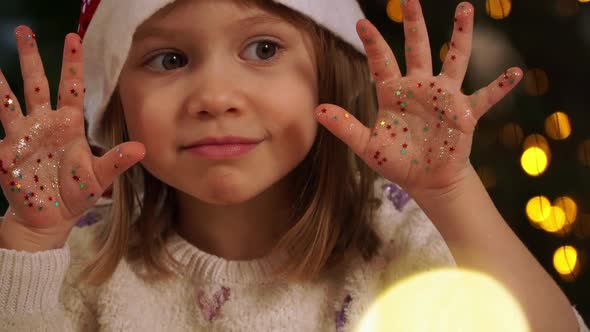 Girl Shows Palms with Glitters and Confetti on Background of Christmas Tree