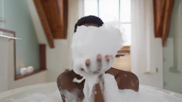Smiling african american attractive woman taking bath and blowing out foam in bathroom alt