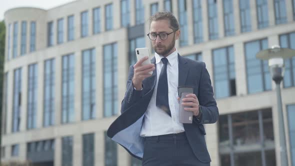 Portrait of Concentrated Nerd Businessman Using Smartphone Outdoors in Front of Office Business alt