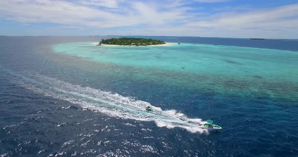Aerial drone view of man and woman on an inflatable tube towing behind a boat to a tropical island alt