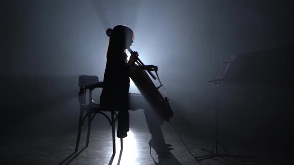Girl Bows the Cello in a Smoky Room at Night. Silhouette. Black Smoke Background alt