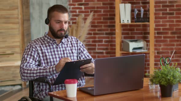 Disabled Person in Headset is Sitting with Clipboard in Front of Table with Laptop Coffee and Office alt