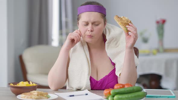Front View of Young Obese Woman Eating Unhealthy Chips and Burger Smelling Tomato Writing Calories alt