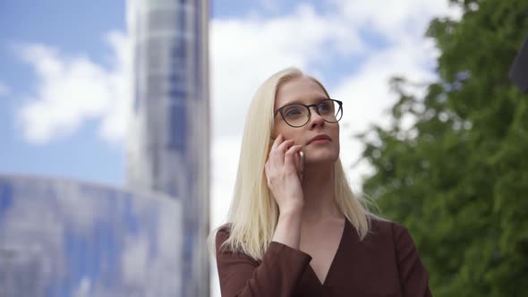 Business Woman Blonde in Glasses is Actively Talking on the Phone Against the Background of High