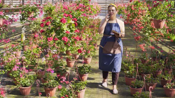 Woman Talks on Smartphone Writing in Notebook in Greenhouse
