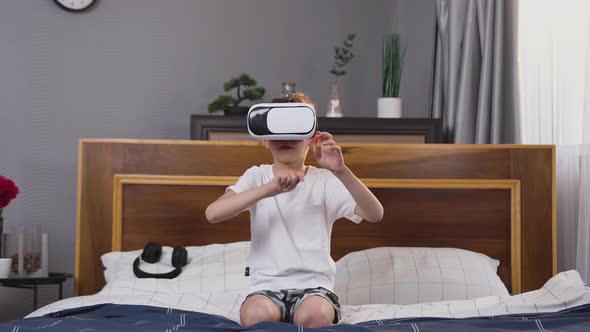 Small Boy in Virtual Reality Headset Sitting on Bed and Playing Game on Imaginary Screen alt