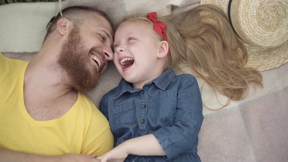 Top View of Laughing Joyful Young Bearded Father and Cute Little Daughter Lying on Blanket Talking alt