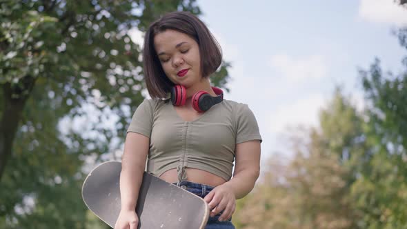 Medium Shot Portrait of Positive Little Woman with Skateboard Gesturing Strength Looking at Camera alt