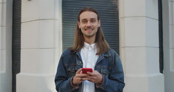 Crop View of Long Haired Guy Raising Head and Looking To Camera While Using His Smartphone. Bearded alt