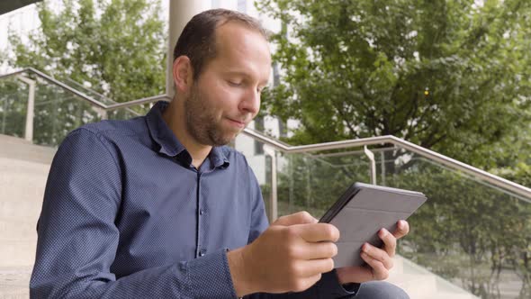 A Caucasian Man Smiles and Nods As He Works on a Tablet and Sits on a Staircase in an Urban Area