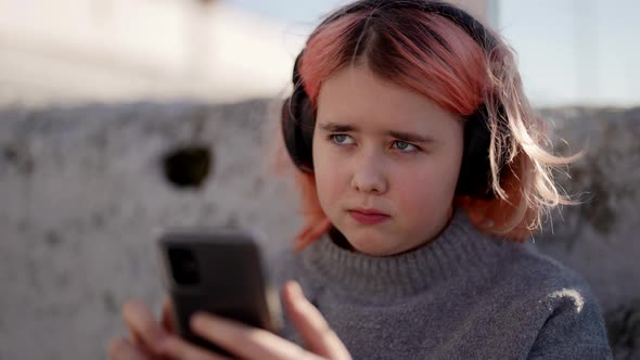 Preteen Girl is Playing in Smartphone and Listening to Music By Wireless Headphones on Street alt
