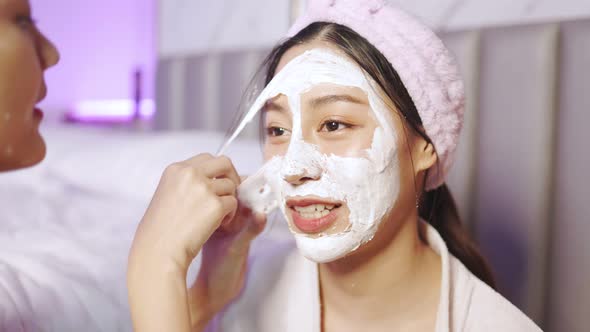 Beautiful young woman in white bathrobe applying a revitalizing  mask her friend's face. alt
