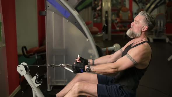 A gray-haired solid man performs exercises for the back and biceps on a training machine alt