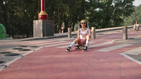 Two Young Girls with Bright Appearance Riding on a City Bridge Sitting on a Skateboard alt