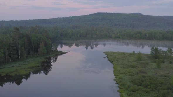 Dawn aerial gliding over flat calm lake covered in thin fog looking over wilderness alt
