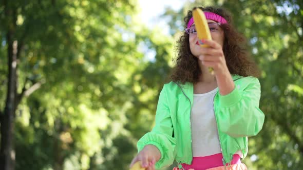 Carefree Laughing Woman in Retro Outfit Imitating Gun Shooting with Bananas Outdoors in Slow Motion alt
