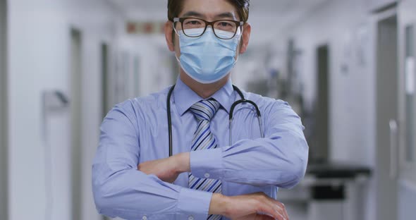 Portrait of caucasian male doctor with arms crossed wearing face mask in the corridor at hospital alt