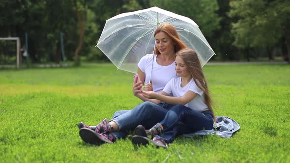Happy mother smiles and plays with her daughter in the park hiding from the rain. alt