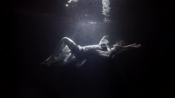 A Beautiful Woman in a Dress Floats on Her Back Under the Water on a Dark Background She Swims To alt
