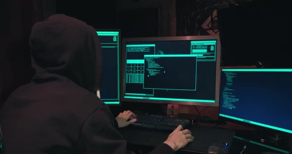 Hacker in a Hood Sits in Front of Computer Screens and Hacks Databases