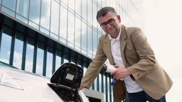 Attractive Older Man Plugging in Charging Cable to to Electric Vehicle alt