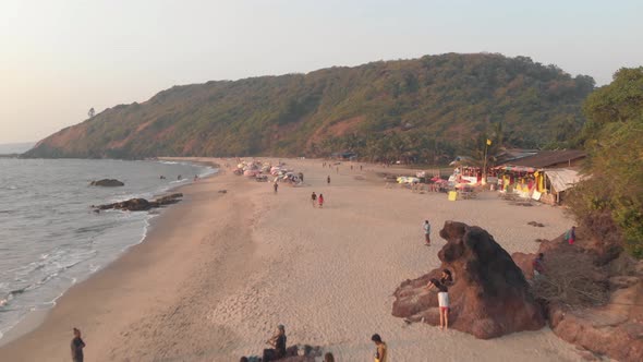 People stroll along Arambol beach, serene ocean waters. Goa, India. Aerial forward alt