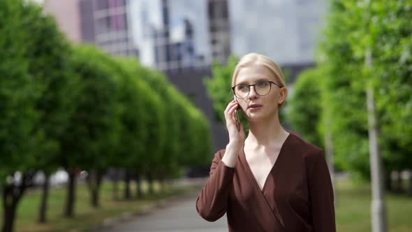 A Serious Confident Woman Answers the Phone As She Walks Down an Alley with Green Trees alt