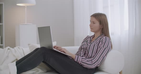 Young Woman Is Browsing Internet, Using Laptop, Sitting on Couch at Home, Working Remotely or alt