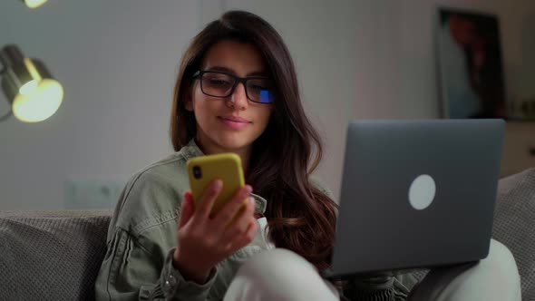 a Girl with Glasses Works on a Laptop at Home on the Couch and Speaks on a Mobile Phone