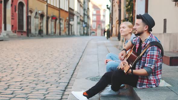 Tourists Sitting on Sidewalk Playing Guitar and Having Rest alt