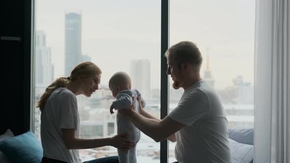 parents teach toddler to walk while sitting by a large window