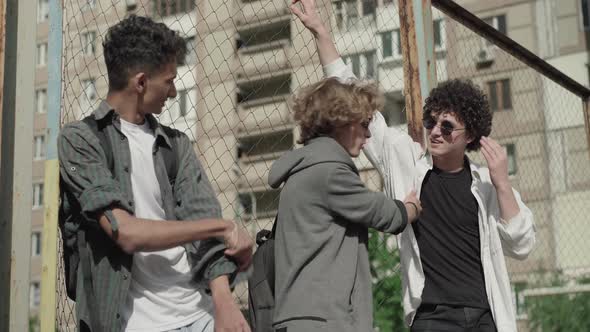 Three Happy Male Friends Standing at Mesh Fence and Talking. Smiling Caucasian and African American alt