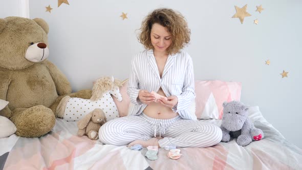 Young Smiling Pregnant Woman Choosing Socks For Future Baby Wearing Pajamas At Home. alt