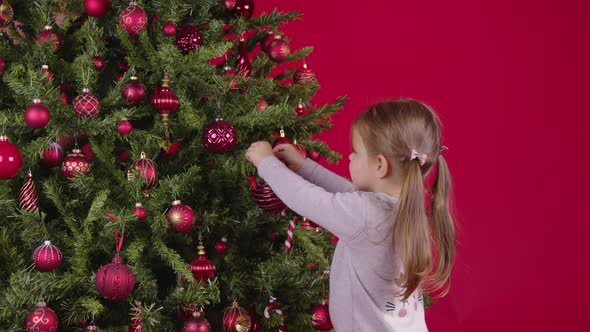 Little Girl Decorating Xmas Tree, Hangs Beautiful Christmas Ball, Red Background