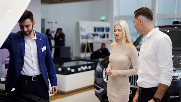Salesman Showing Car Engine Under Open Hood to Couple in Auto Showroom alt