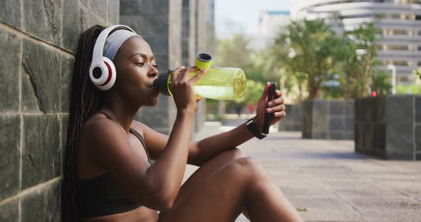 African american woman exercising outdoors drinking water and using smartphone in the city alt