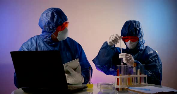 People in Protective Clothing with Hoods, Respirators, Glasses and Gloves Sit at a Table with a alt
