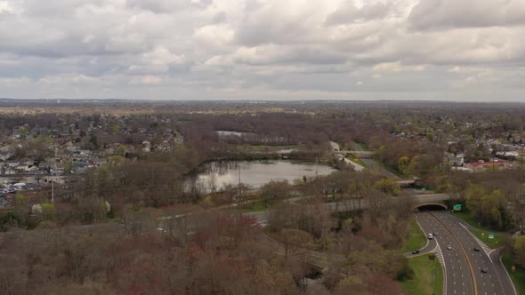 A drone view over a lake, surrounded by dry trees and paved roads. It is a cloudy day and the water alt