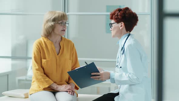 Elderly Woman and Female Doctor Talking on Medical Couch in Clinic alt